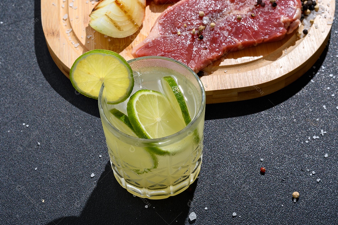 Tábua de madeira com carne temperada e copo de caipirinha